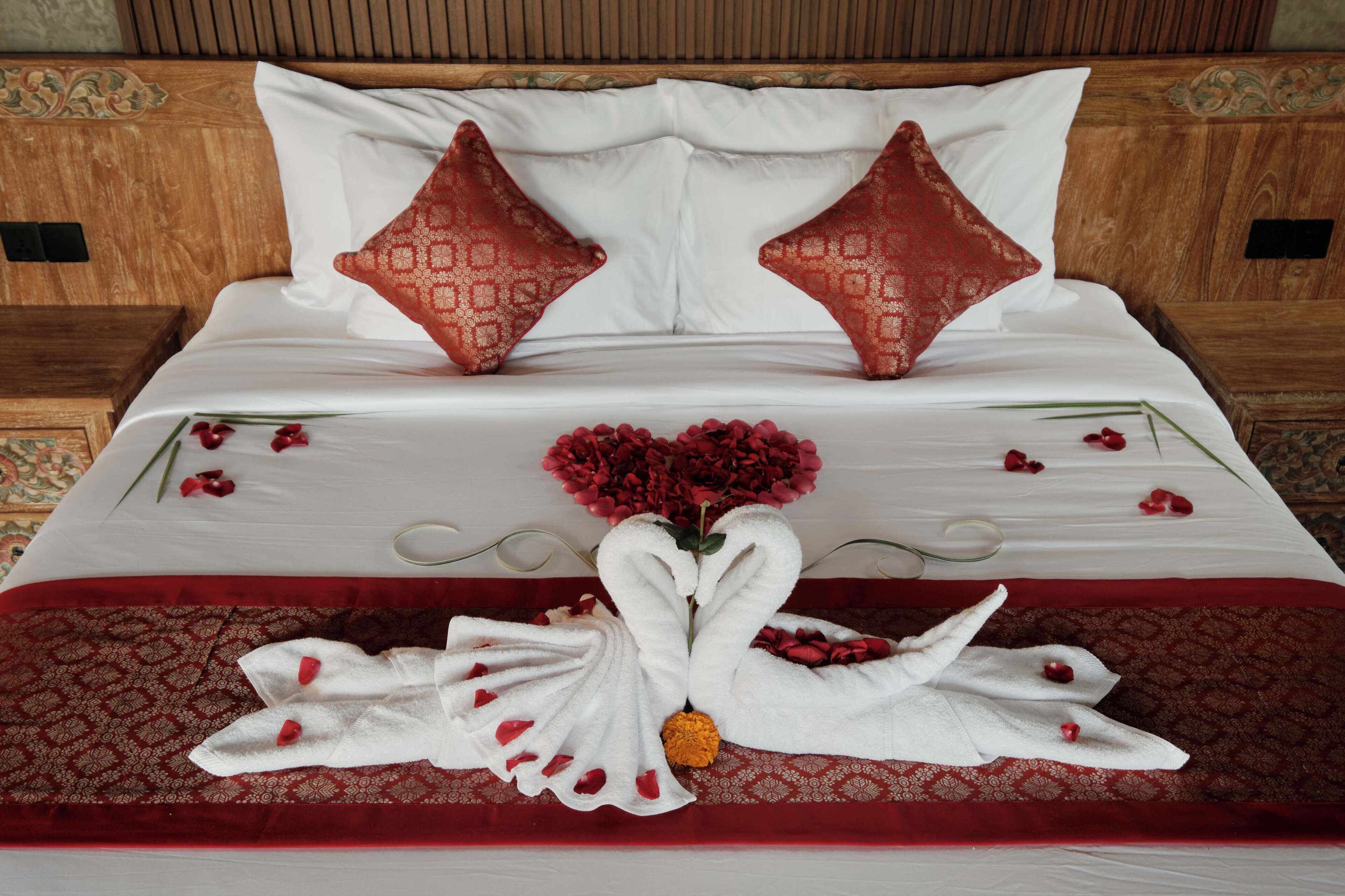 A neatly made bed with a red and white patterned bedspread, two decorative pillows, and a towel arrangement in the shape of swans with a flower centerpiece.