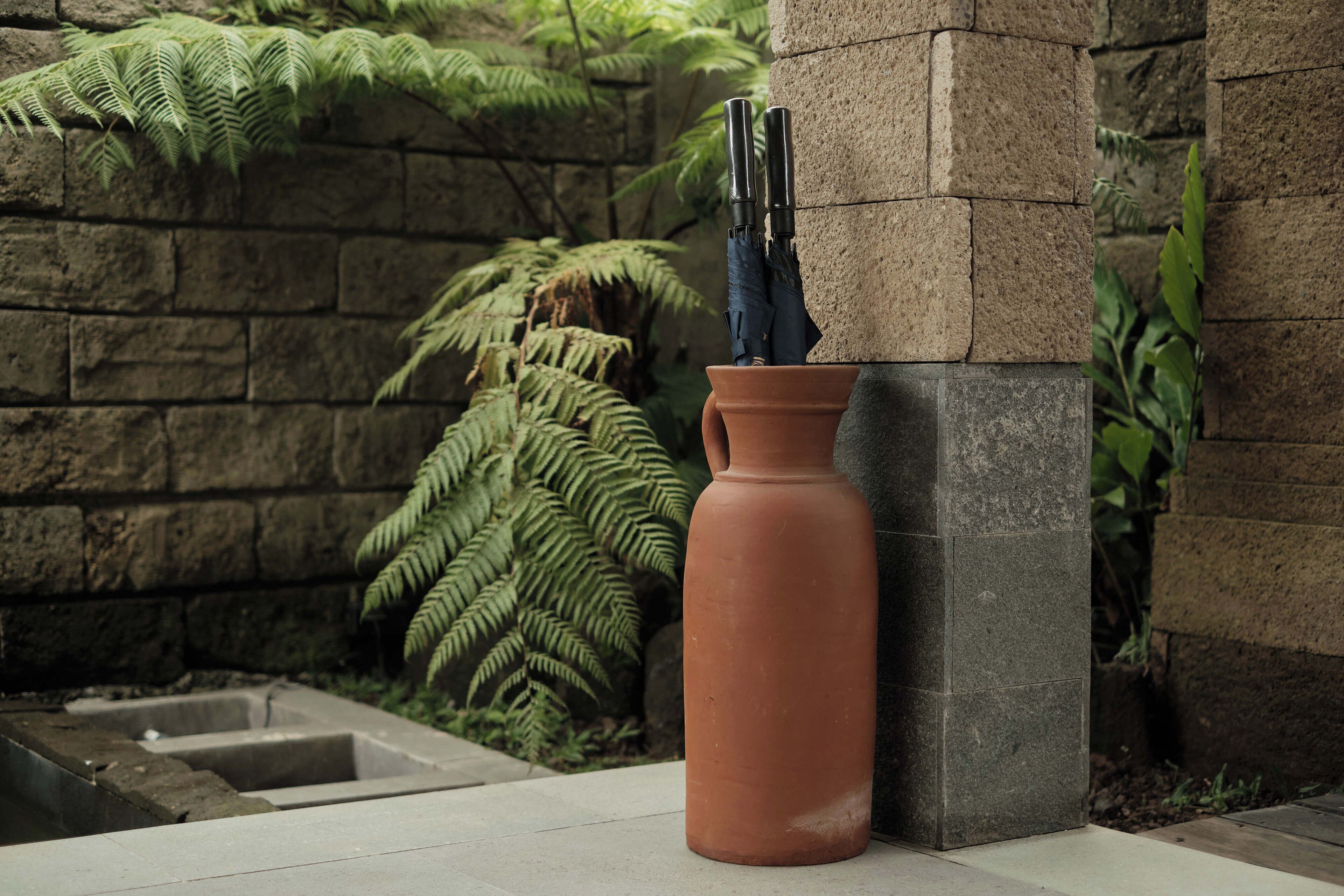 A terracotta vase with a pair of black umbrellas on a stone pillar in a garden setting.