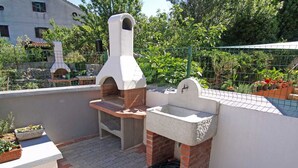 Ein Außenküchenbereich mit Pizzaofen und Spüle, umgeben von einem Garten, mit einem Gebäude im Hintergrund.