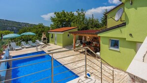 Area piscina con poltronne da relax e ombrelloni, zona barbecue e vista su un edificio verde con parabola.