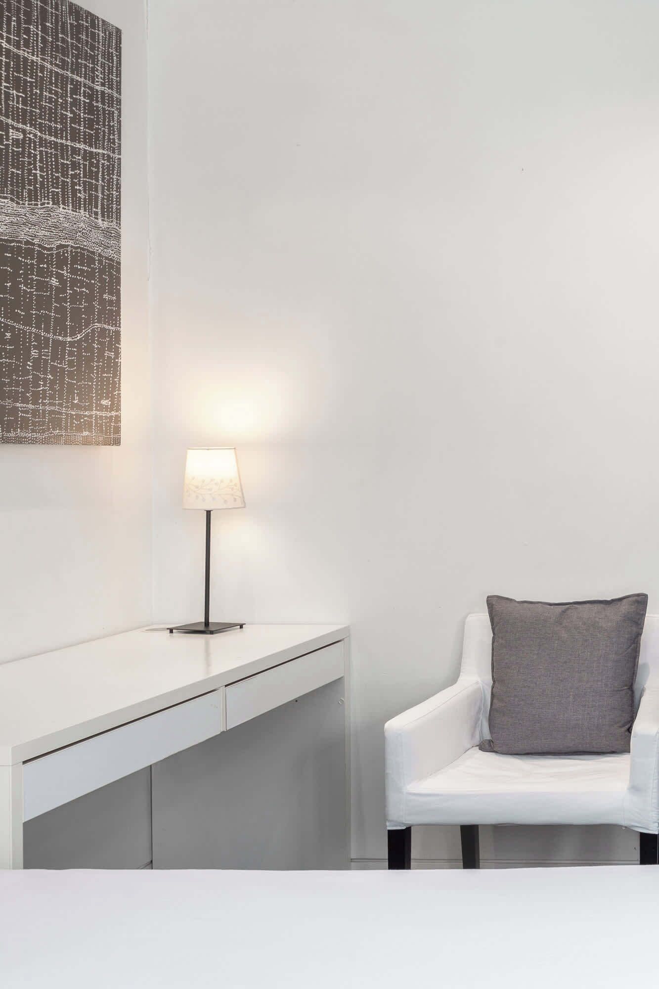 Une pièce au décor minimaliste, avec un bureau blanc, une lampe et une chaise agrémentée d’un coussin gris.