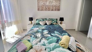 Ein Schlafzimmer mit floralem Bettüberwurf, zwei Nachttischen inklusive Lampen, einem Gemälde an der Wand und Blick ins Freie durch transparente Vorhänge.