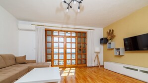 Un soggiorno moderno con un divano, un tavolino da caffè bianco e un pavimento in legno. È presente un'ampia porta-finestra scorrevole che dà su un balcone, e un televisore montato a parete.