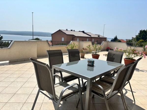 Une terrasse avec une table et des chaises, des plantes en pot et une vue sur la mer et les bâtiments.