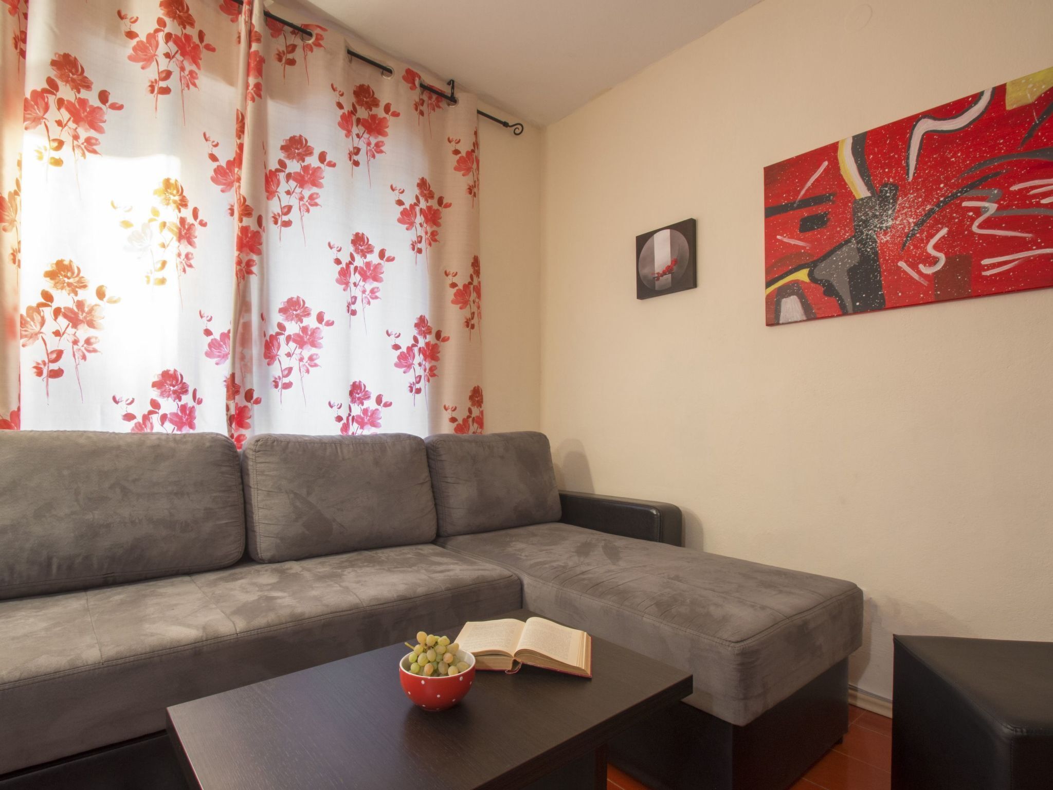 Ein Wohnzimmer mit einem grauen Sofa, einem Couchtisch mit Obstschale und offenem Buch, einem roten und weißen Blumenvorhang und zwei Bildern an der Wand.