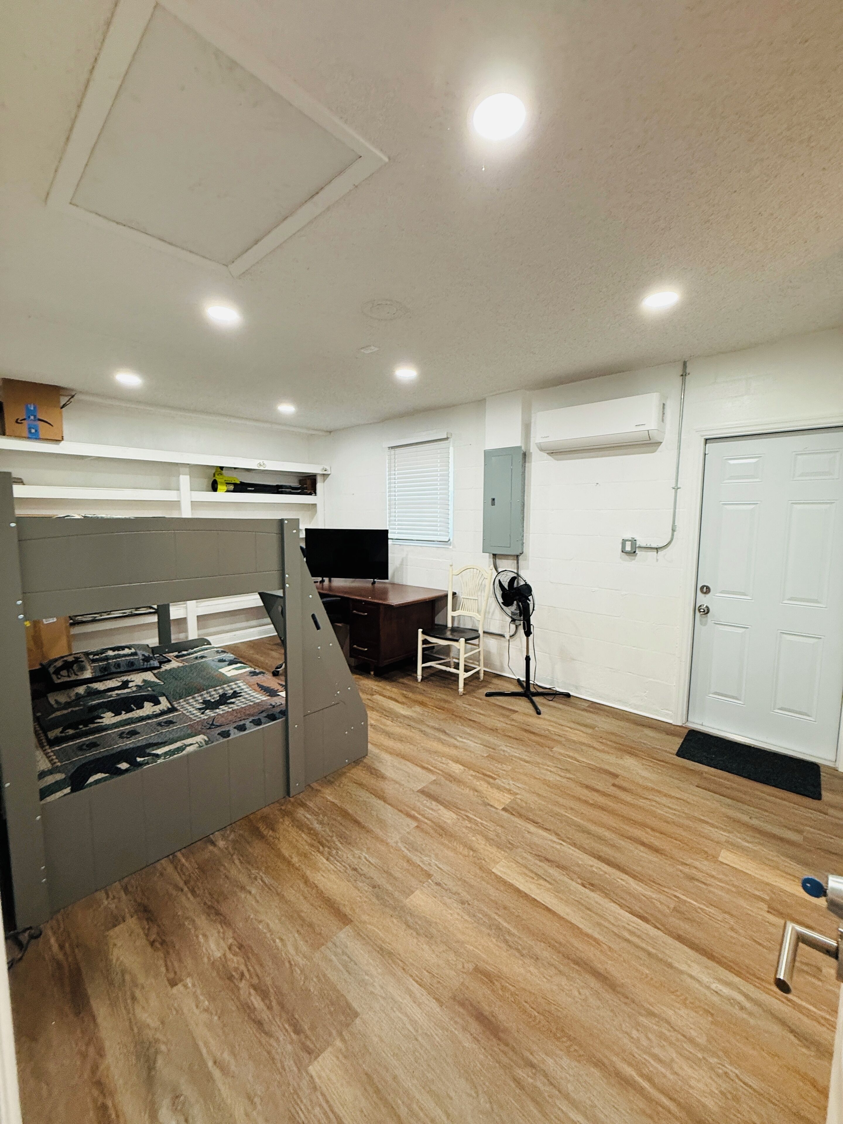 A room with a bunk bed, a desk, a chair, a fan, and an air conditioning unit.