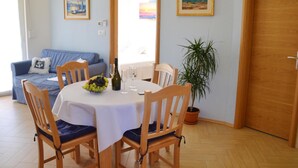Zona pranzo con un tavolo rotondo, sedie, una bottiglia di vino e una ciotola di frutta. Sullo sfondo, un divano blu con una coperta a quadri, una pianta in vaso e due quadri appesi al muro.