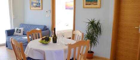 A dining area with a round table, chairs, a bottle of wine, and a bowl of fruit. There is a blue sofa with a checkered cover in the background, a potted plant, and two framed pictures on the wall.