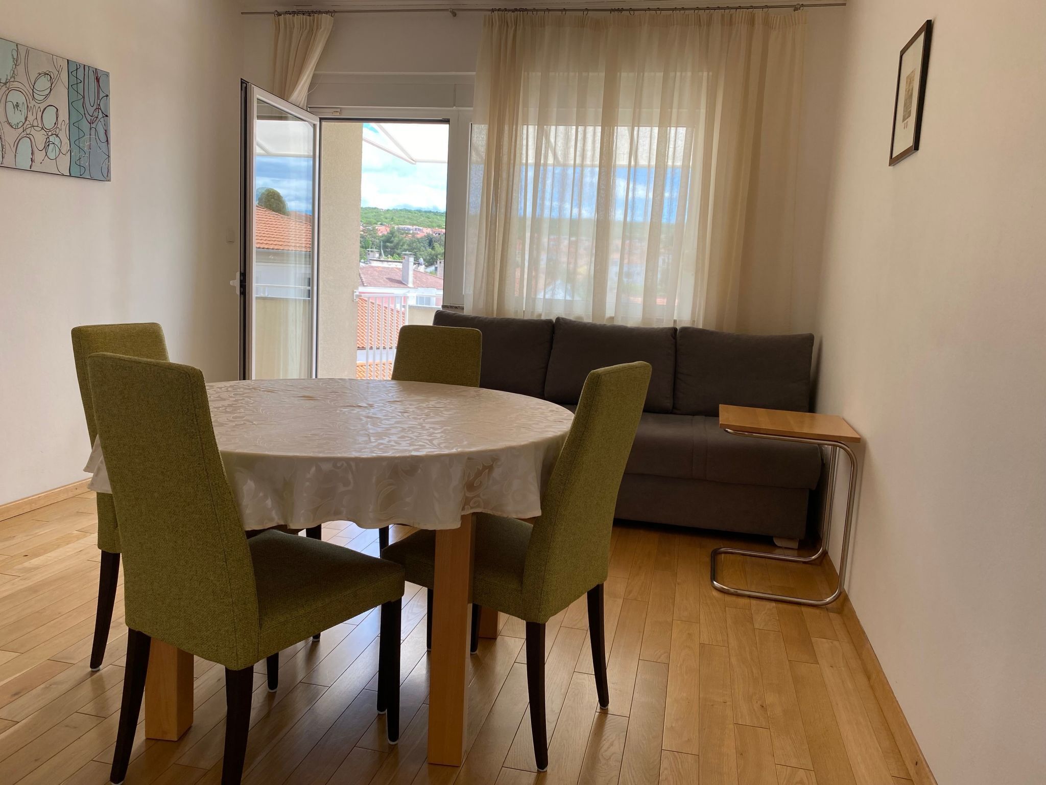 A dining area with a round table, four chairs, a sofa, and a view of a balcony with a railing.