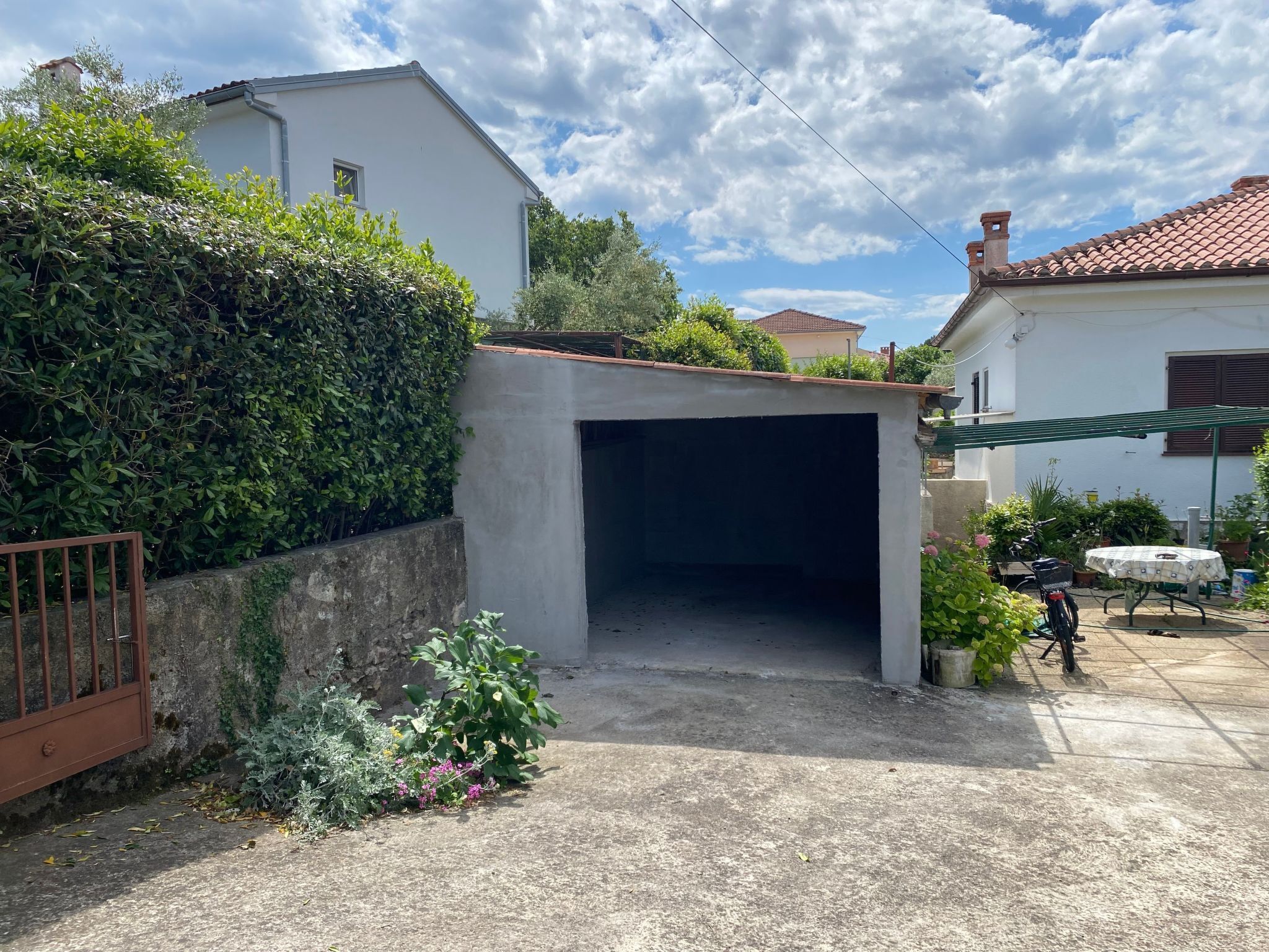 Un garaje residencial con entrada de hormigón, rodeado de follaje y un tejado de tejas.