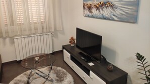 A living room with a TV on a dark wooden stand, a glass coffee table, a white radiator, and a painting of a lighthouse on the wall. - Große Wohnung in Gabonjin mit Großem Garten by Interhome (Krk/Milohnići)