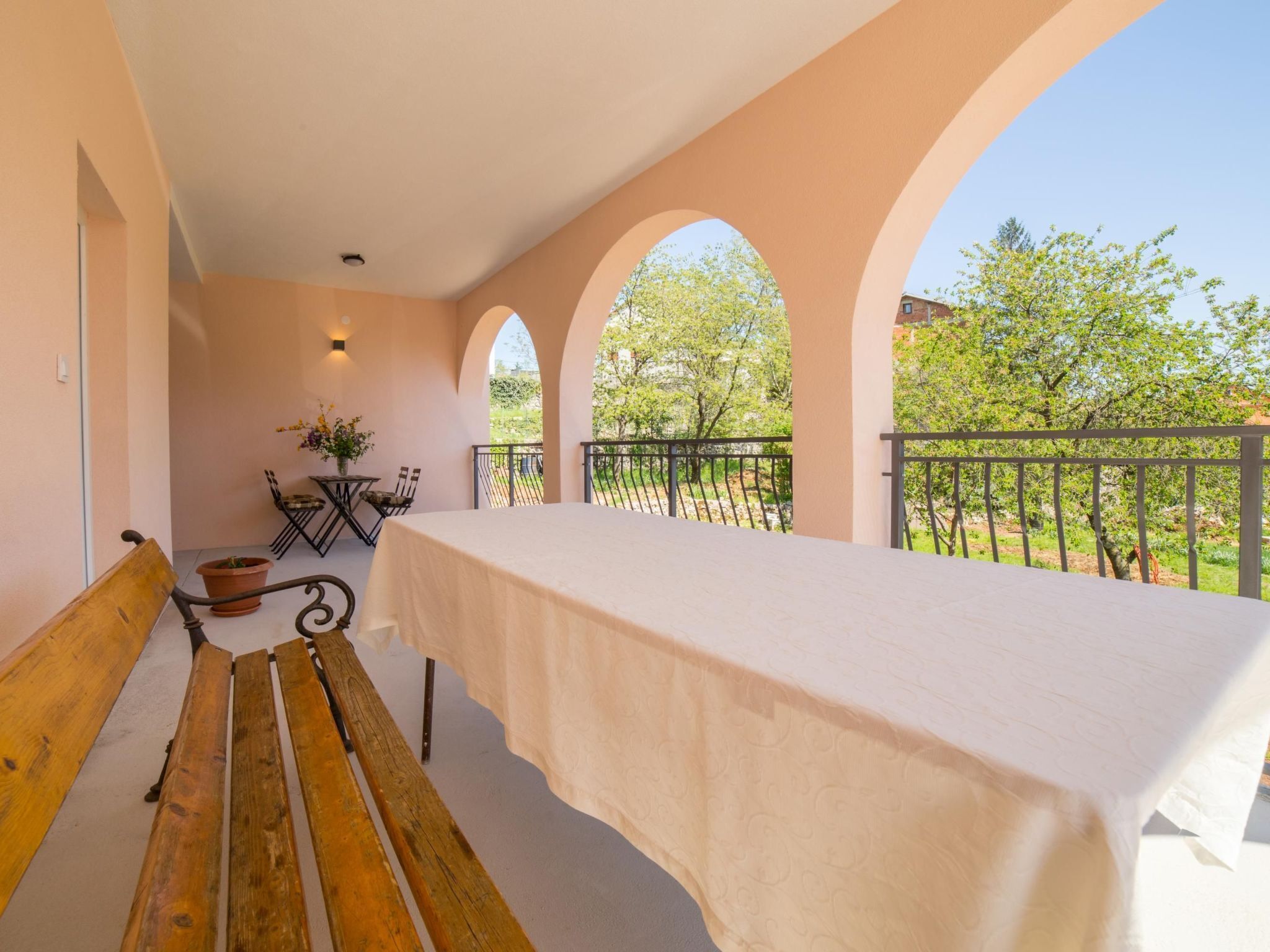 Ein Balkon mit einer Holzbank, einem Tisch mit weißem Tischlein und Blick auf Bäume und Gebäude.