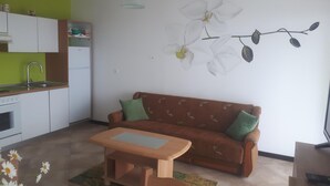 Een knus woongedeelte met een kitchenette, een bank met kussens, een klein houten tafeltje en een orchidee schilderij aan de muur.