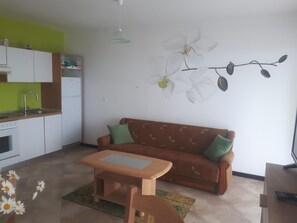 A compact living space with a kitchenette, a sofa with cushions, a small wooden table, and a wall-mounted orchid painting. - Gemütliche Ferienwohnung mit toller Aussicht by Interhome (Lovran)