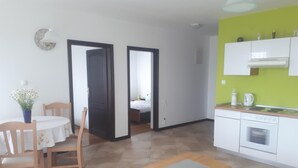 Een kleine keuken met witte kasten en een groene accentmuur, een eethoek met een tafel en stoelen, en een zichtbare slaapkamer door een geopende deur.