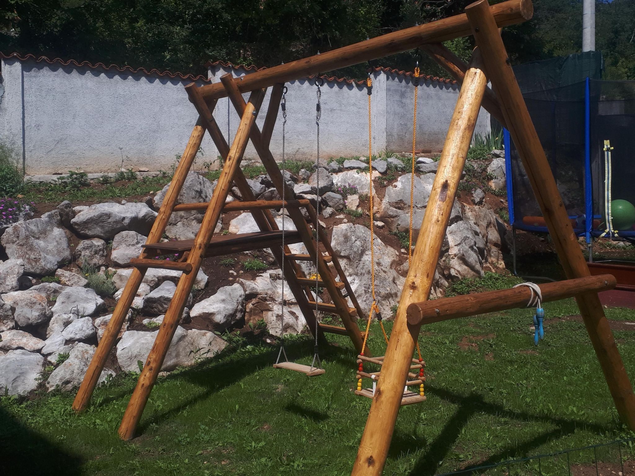 Un conjunto de columpios de madera en un jardín con un muro de piedra y una valla azul al fondo.