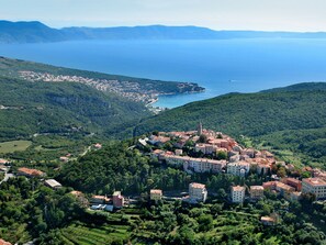 A coastal town with a central cluster of buildings surrounded by greenery, a clear view of the sea, and a hilly landscape in the background.