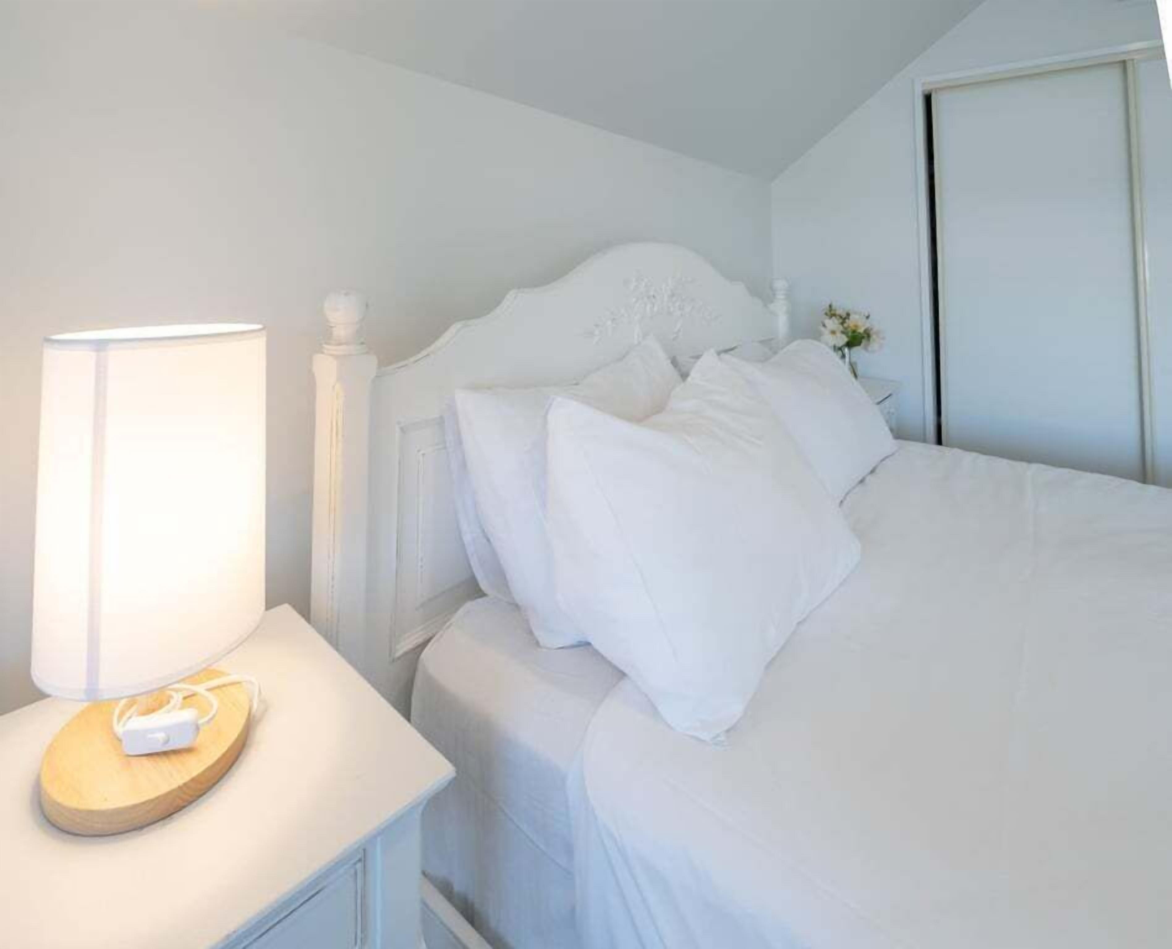 Un letto rifatto con biancheria e cuscini bianchi, un comodino con una lampada e un piccolo vaso con fiori.