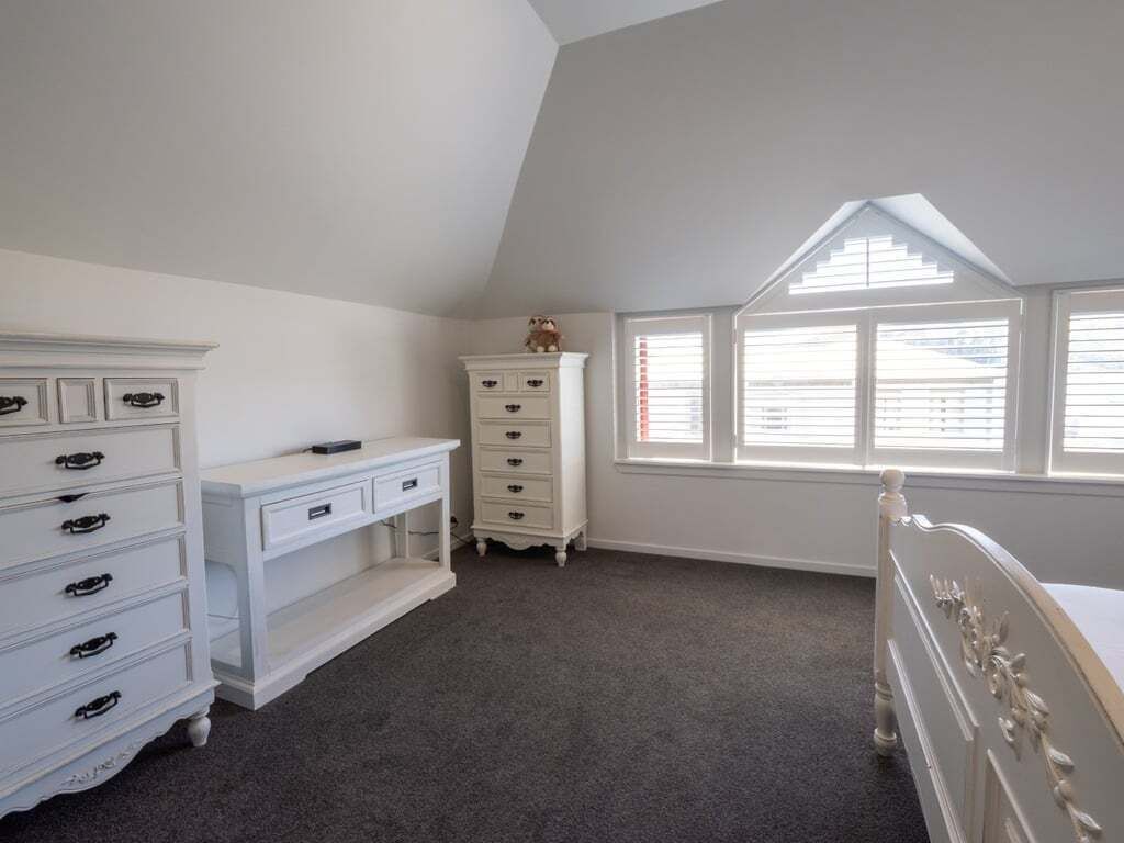 Un dormitorio con techo inclinado, muebles blancos, una cama y una ventana con persianas.