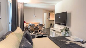 Een moderne hotelkamer met een bed, een zithoek met rieten stoelen, een aan de muur bevestigde televisie en een tafeltje met een plant.