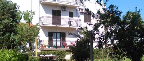 A multi-story building with balconies, surrounded by greenery and a low wall.