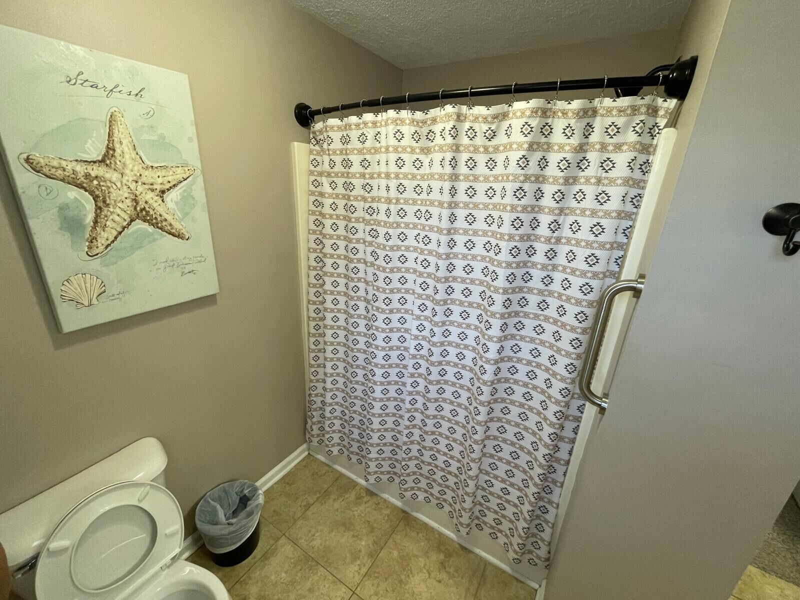 A bathroom with a shower curtain, a toilet, a trash bin, and a wall-mounted starfish artwork.