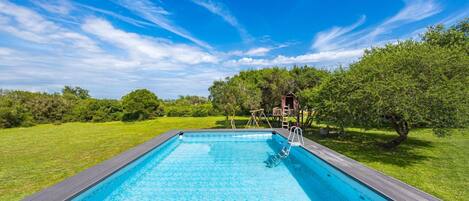 Une piscine rectangulaire dotée d’une terrasse en bois, entourée d’une pelouse verdoyante et d’arbres.