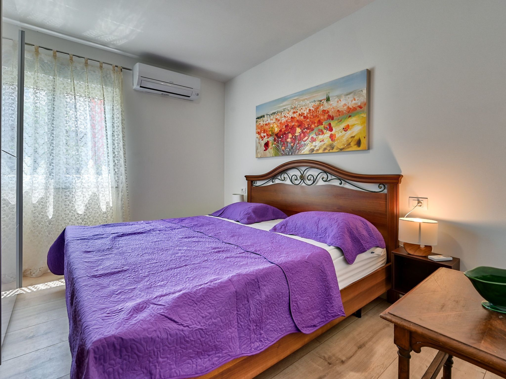 A bedroom with a bed, a painting, an air conditioner, a lamp, and a table.