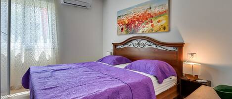 A bedroom with a bed, a painting, an air conditioner, a lamp, and a table.