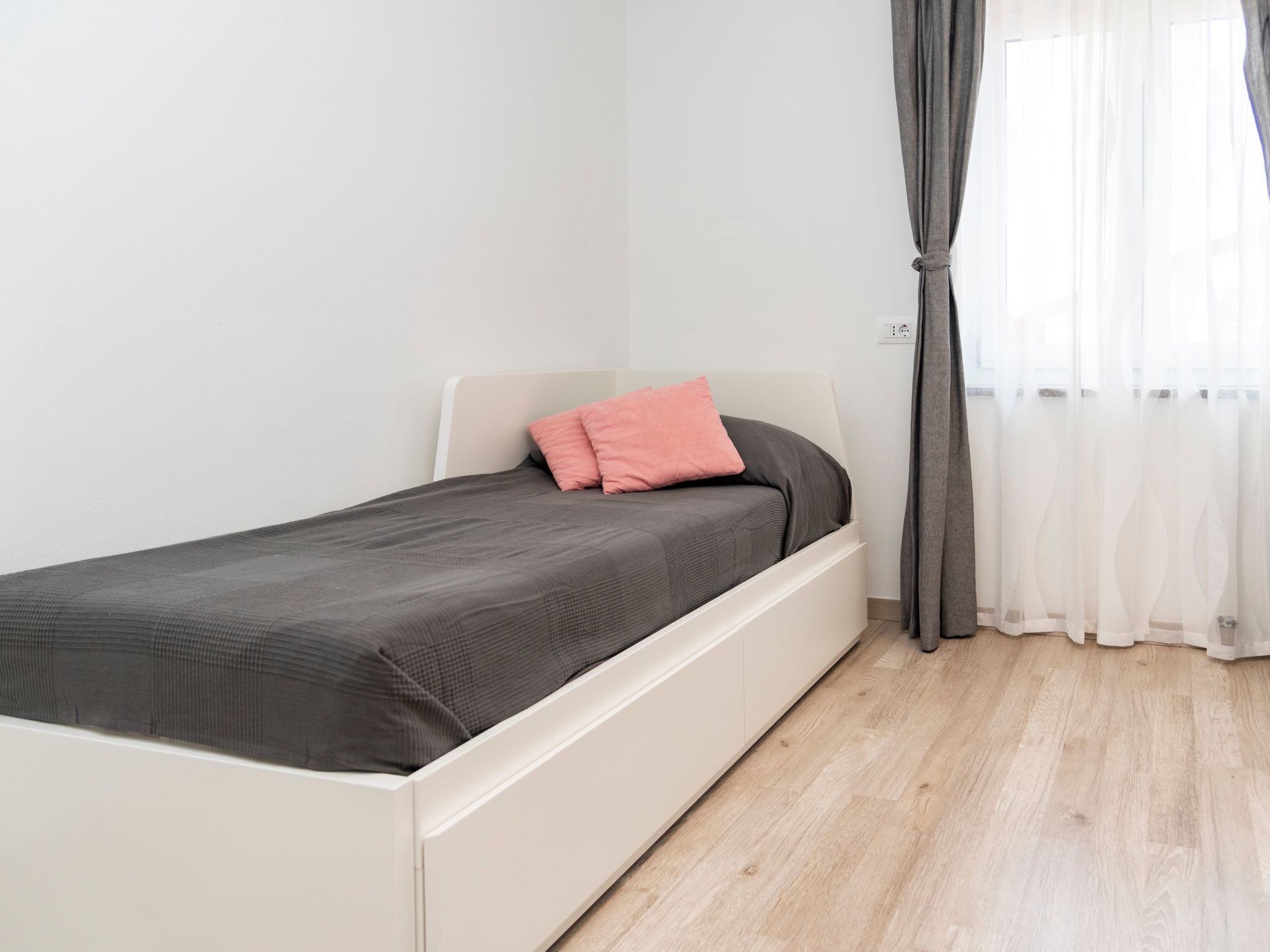 A single bed with a grey mattress and a pink pillow in a room with white walls and a wooden floor.