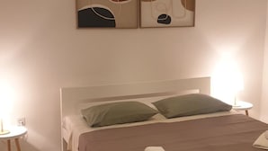 A bedroom with a white bed, green pillows, a brown blanket, a white towel, a bedside table with a lamp, and abstract wall art. - Wohnung a by Interhome (Fažana)