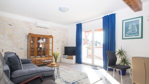 Ein modernes Wohnzimmer mit einer Steinwand, einer Holzkommode, einem Fernseher, einem Sofa, einem Sessel und einem großen Fenster mit Vorhängen.