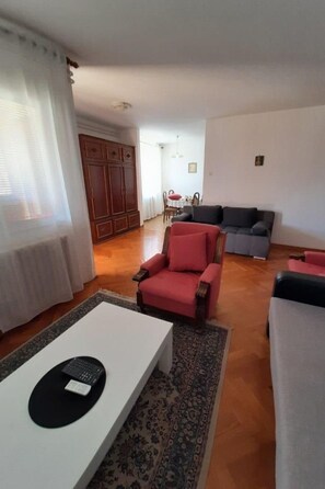 Ein Wohnzimmer mit einem roten Sessel, einem grauen Sofa und einem weißen Couchtisch. Im Hintergrund ist ein Holzschrank und ein Essbereich zu sehen.