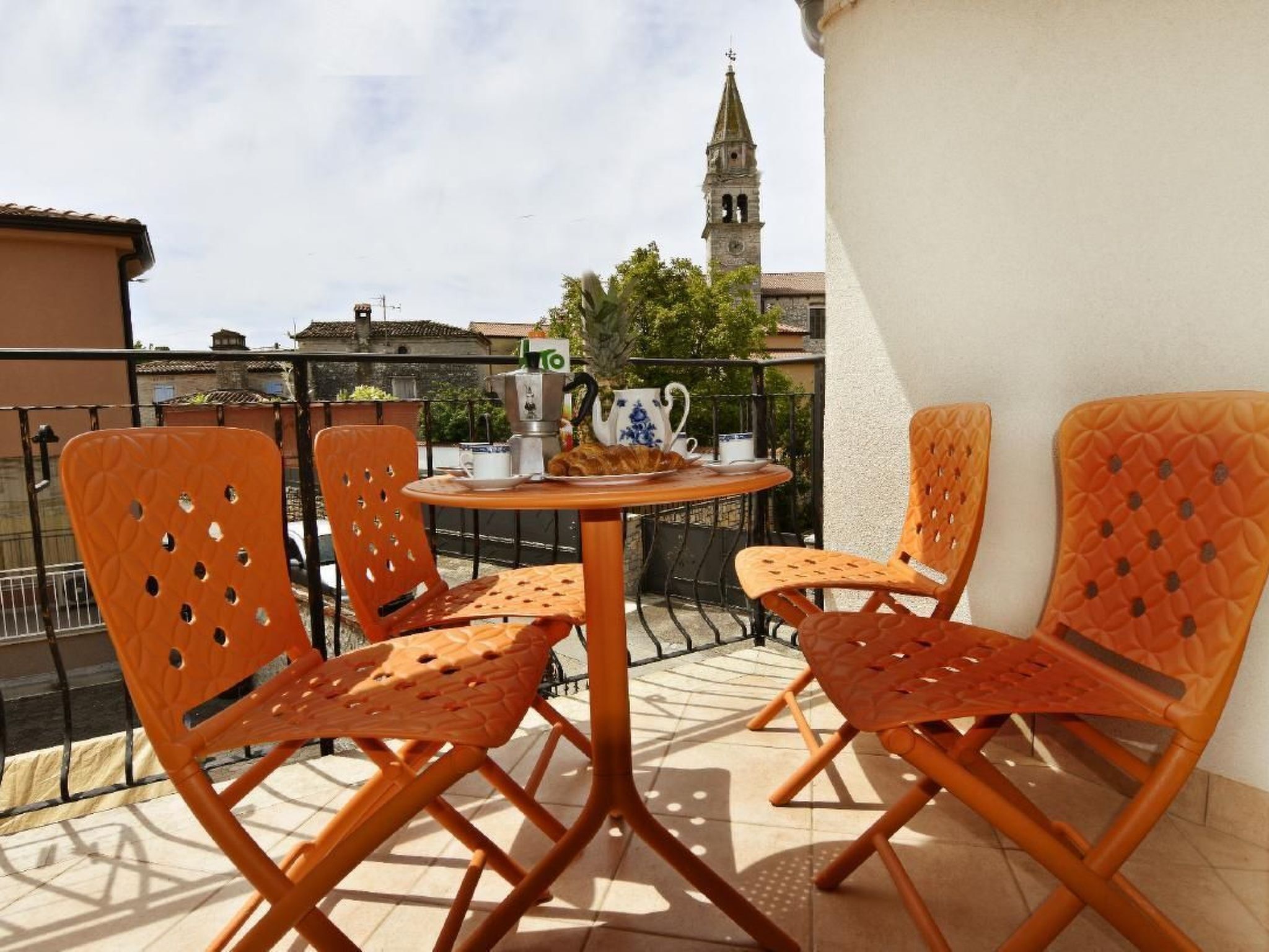 Um terraço com cadeiras laranja e uma mesa, uma máquina de café e um bule. Uma torre do relógio é visível ao fundo.
