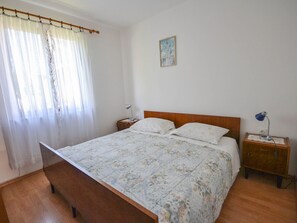 Un dormitori amb un llit de fusta, una tauleta de nit amb una làmpada, una finestra amb cortines lleugeres i un quadre a la paret.