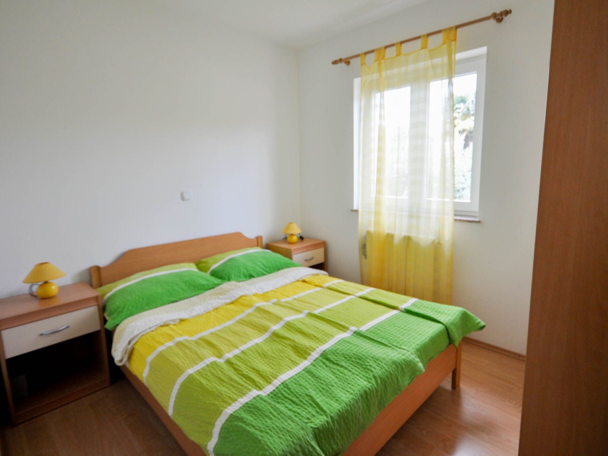Une chambre à coucher avec un lit en bois, une literie à rayures vertes et jaunes, une table de chevet avec une lampe et une fenêtre avec des rideaux jaunes.