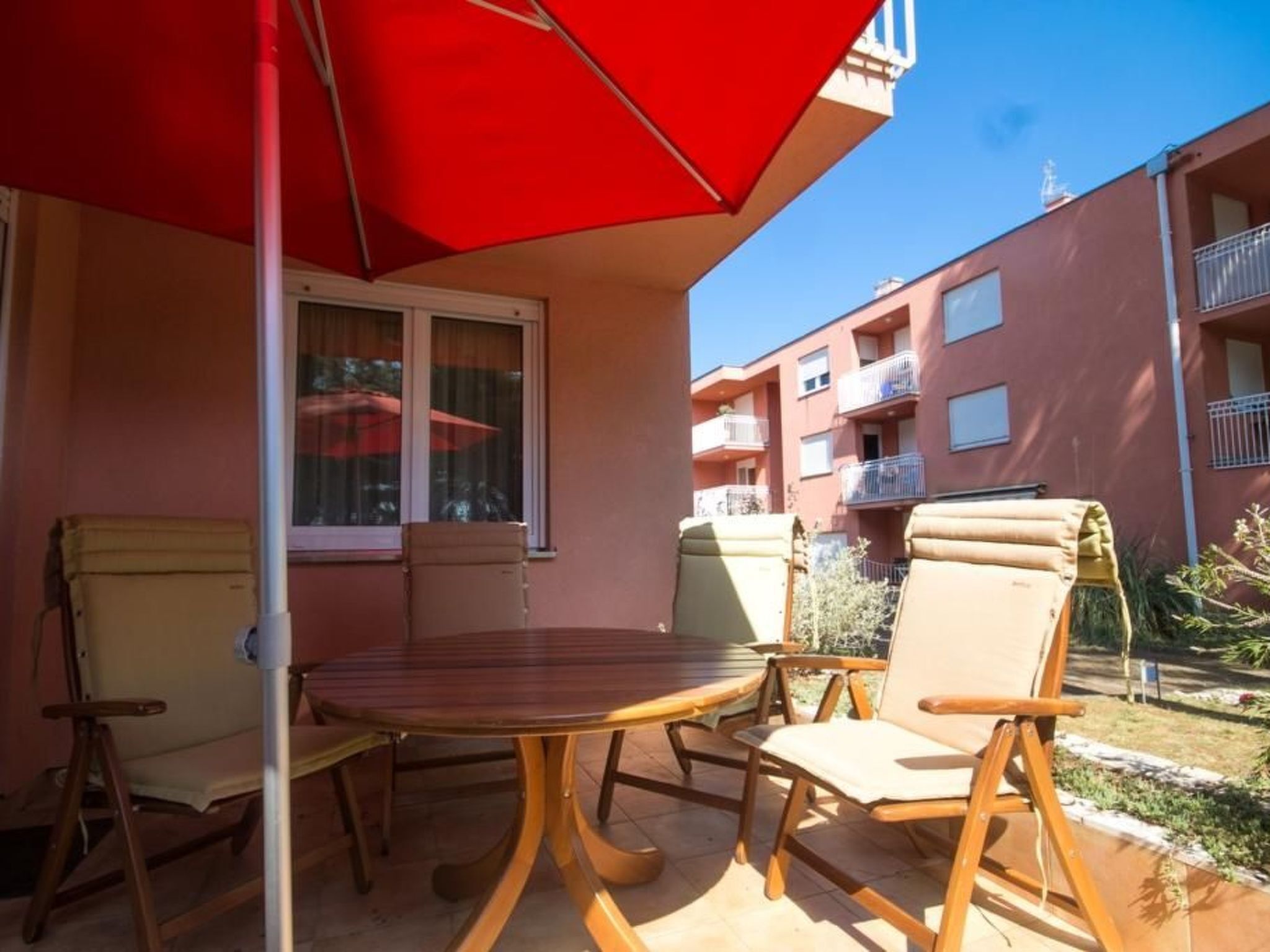 Une terrasse extérieure avec une table ronde et quatre chaises, un parasol rouge et une fenêtre donnant sur un immeuble à plusieurs étages.