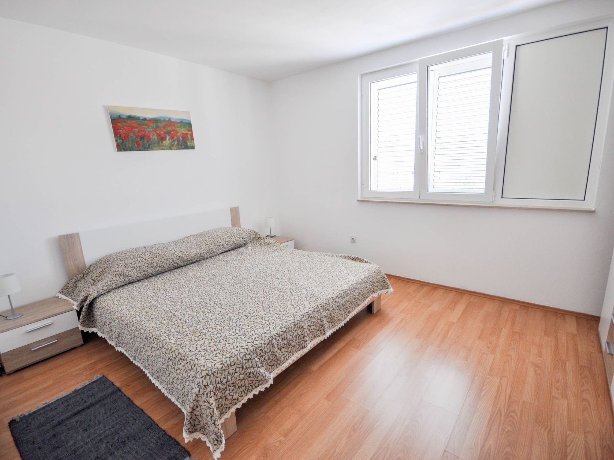 Ein Schlafzimmer mit Holzboden, einem Bett mit gemustertem Überwurf, einem Nachttisch, einer Lampe und einem Fenster mit Jalousien.