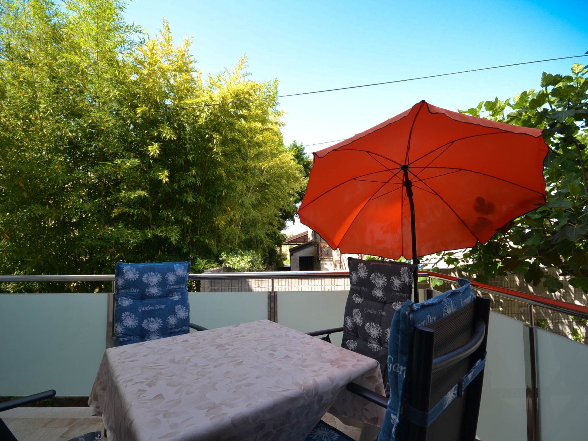 Een balkon met een tafel en stoelen, een oranje parasol en uitzicht op bomen.