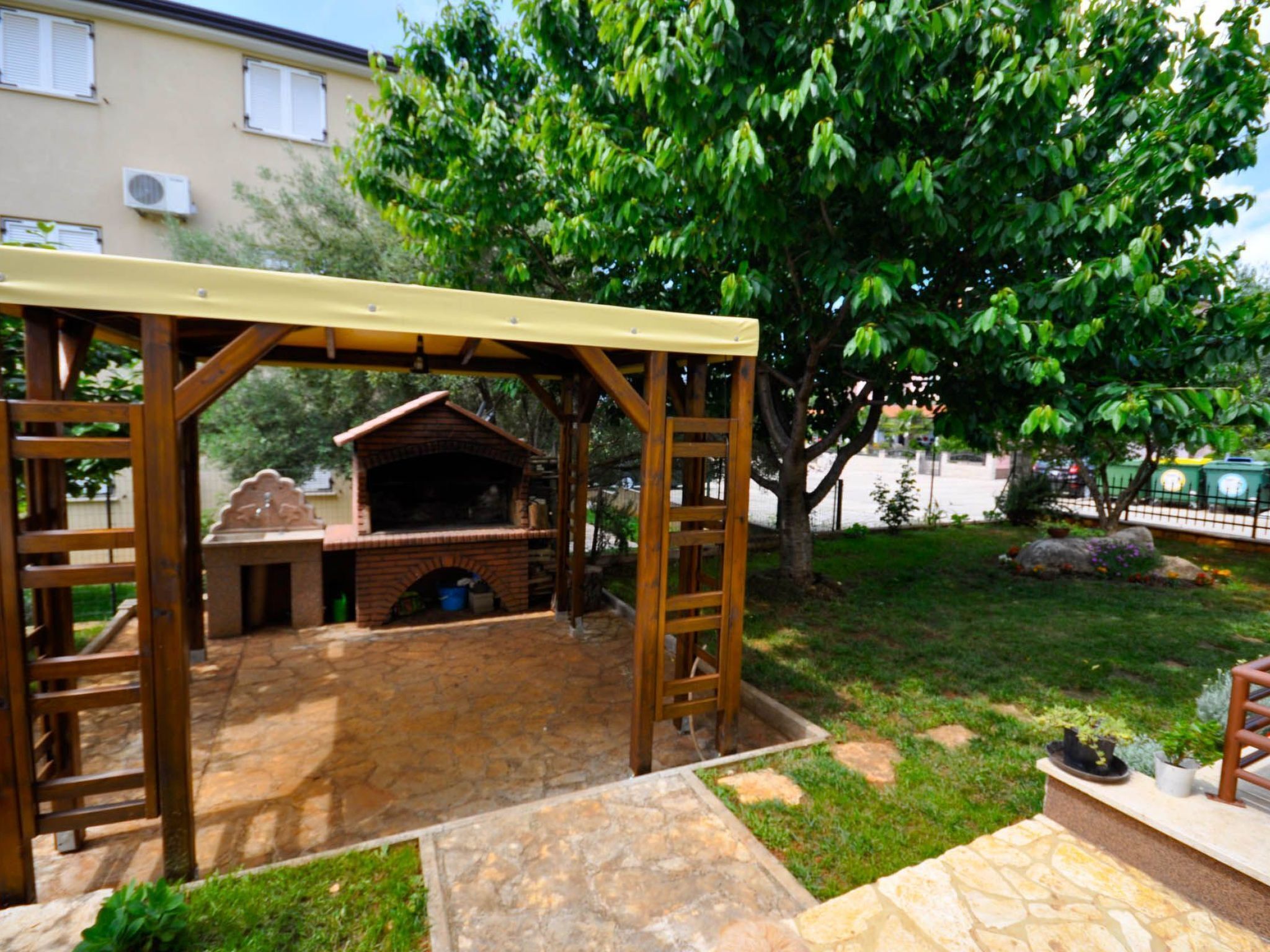 A wooden pergola with a brick barbecue area, stone flooring, and a grassy yard with a tree.
