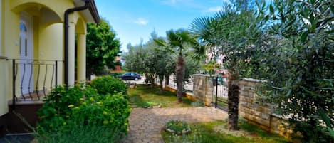 A paved pathway leading to a yellow building with a balcony, surrounded by greenery and a stone wall.