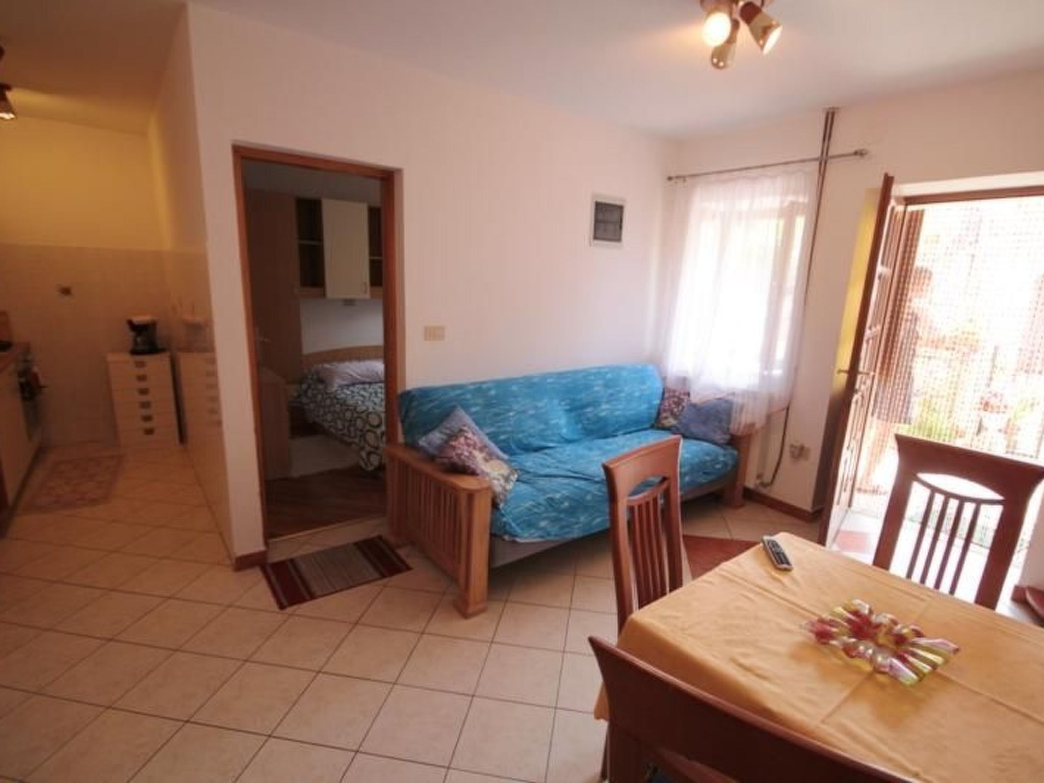 A compact living space with a blue sofa, wooden dining table, and a kitchen area visible through an open doorway.