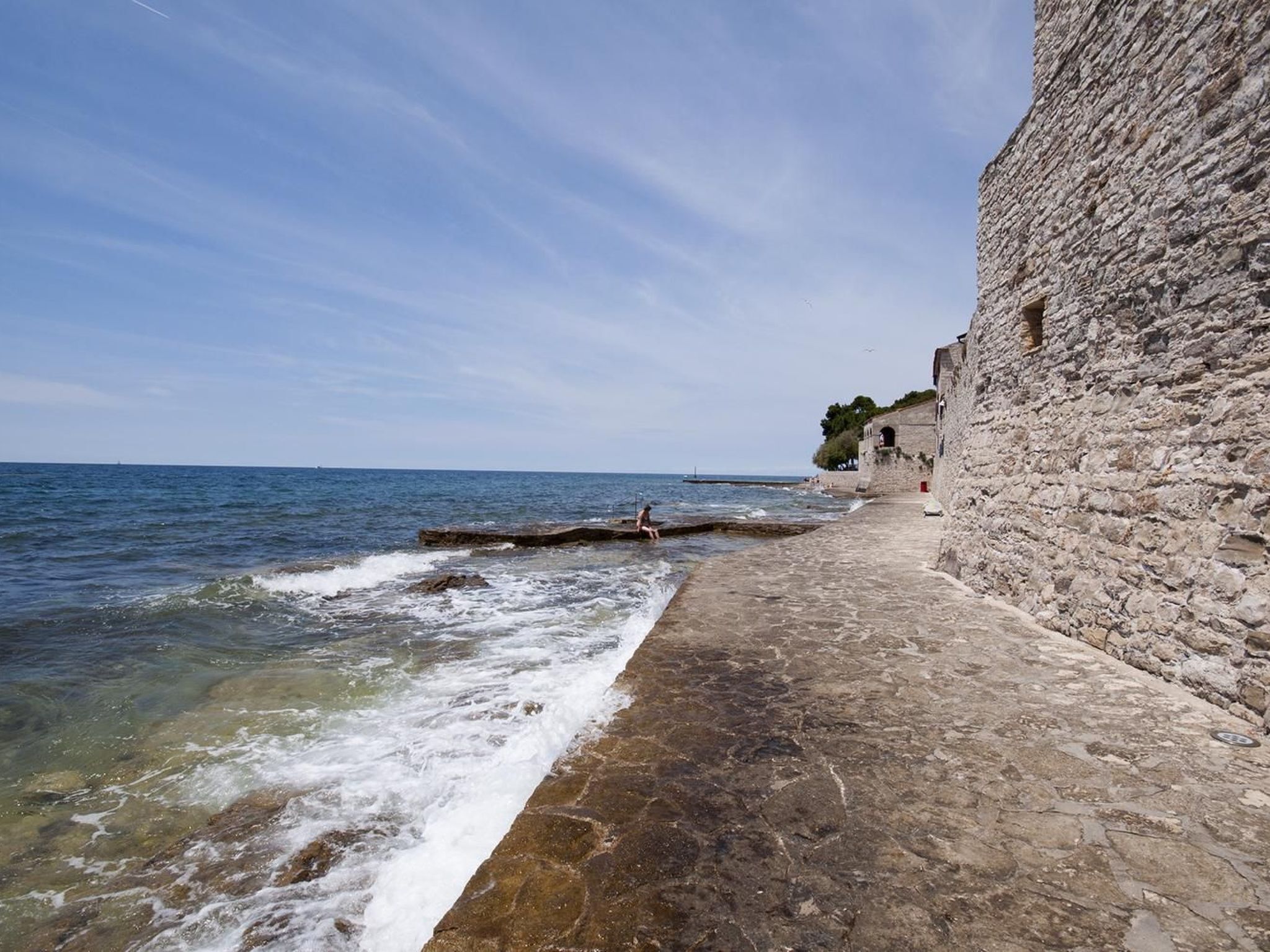 A stone pathway along a coastal area with a clear view of the sea and a stone wall on one side.