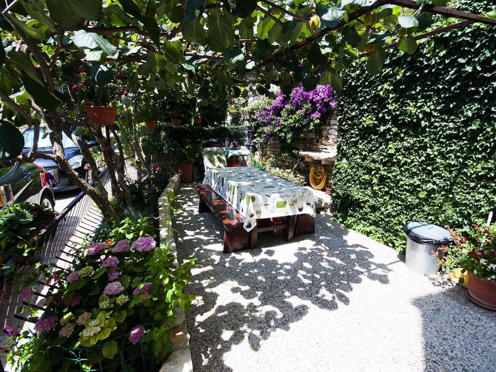 A garden patio with a table and bench, surrounded by lush greenery and flowering plants.