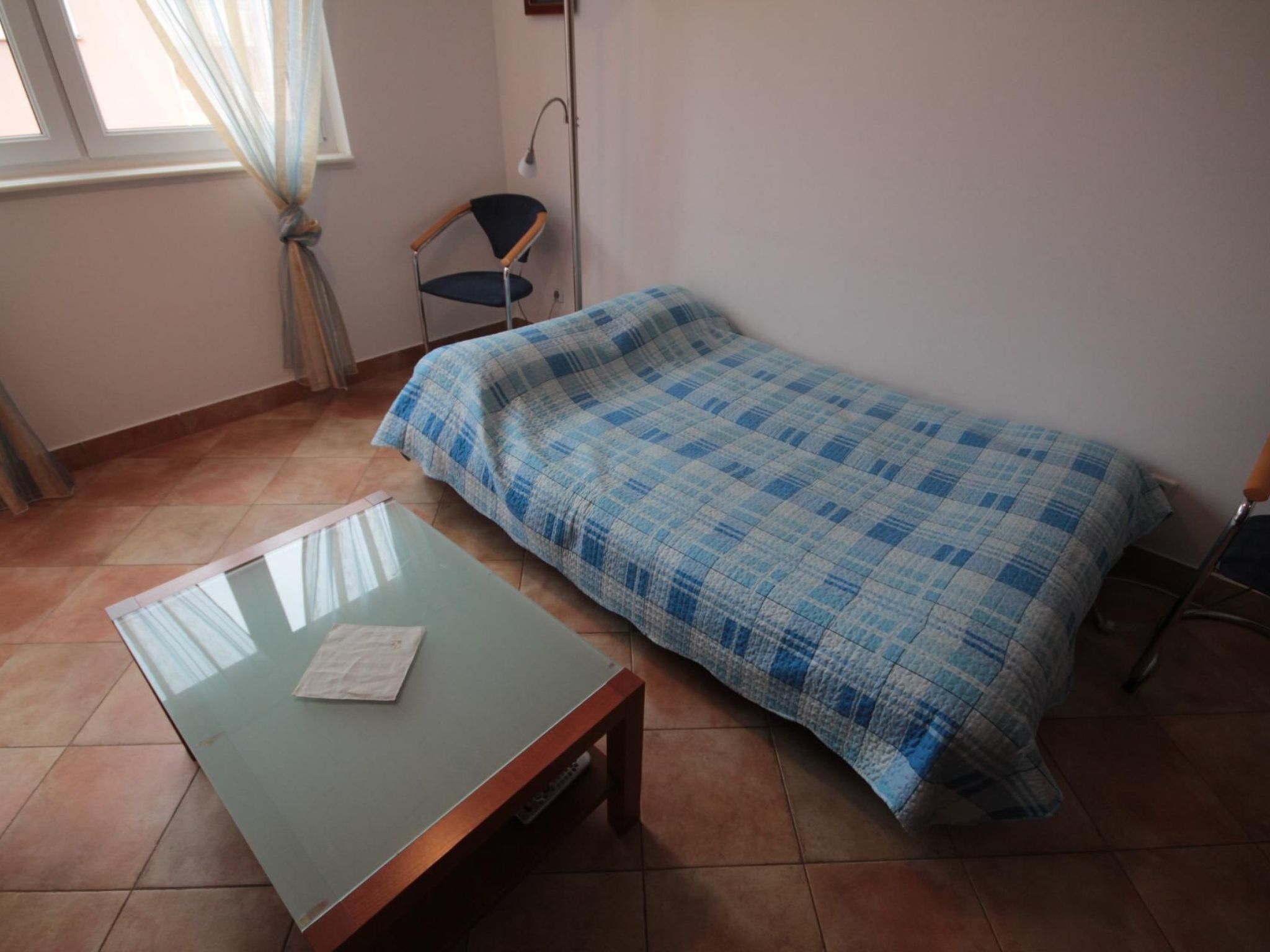 Une petite pièce comprenant un lit simple, une chaise, une table et une fenêtre avec des rideaux.