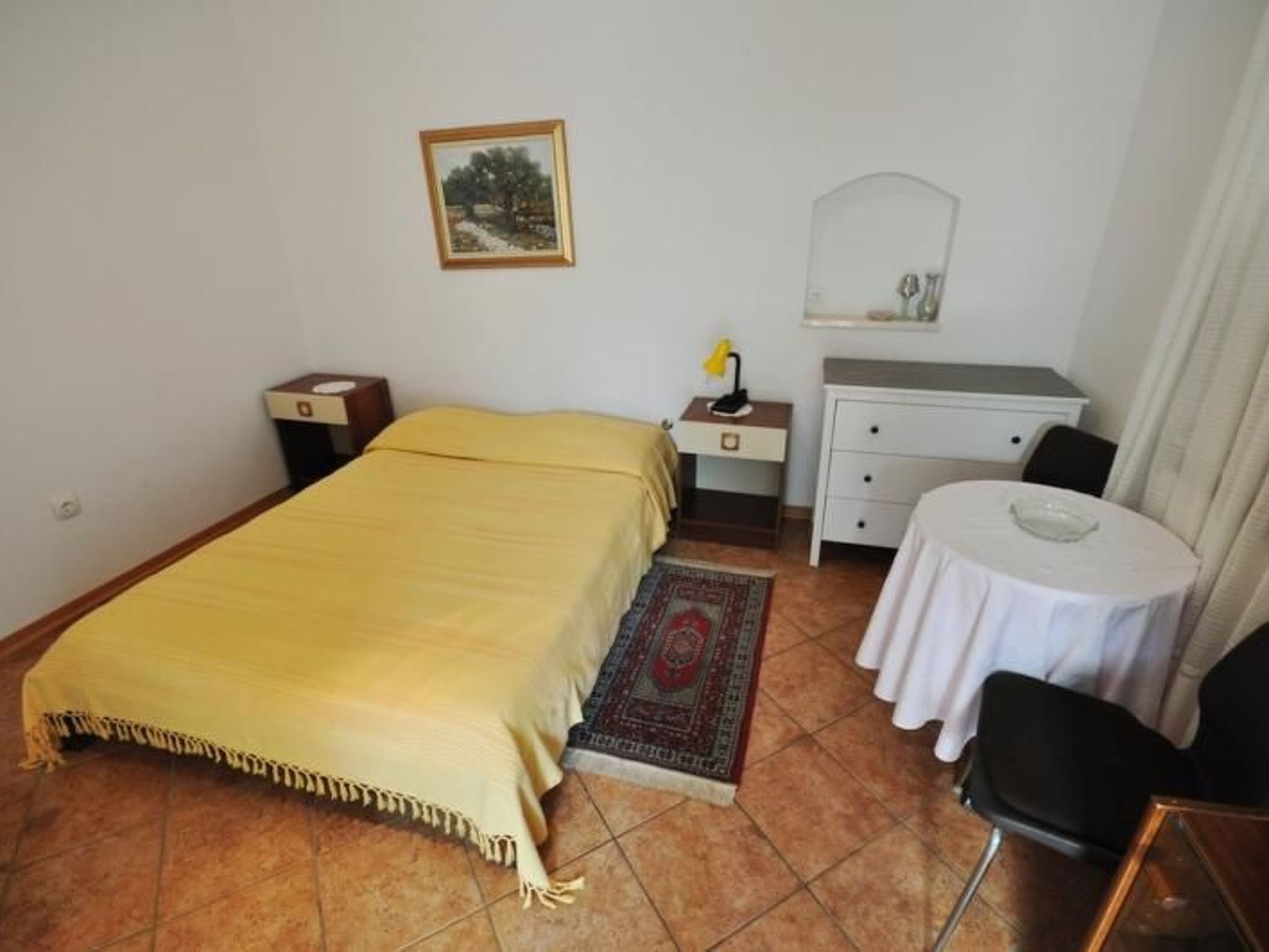 A bedroom with a bed, bedside tables, a desk, a chair, a small table, a rug, a painting, and a mirror.