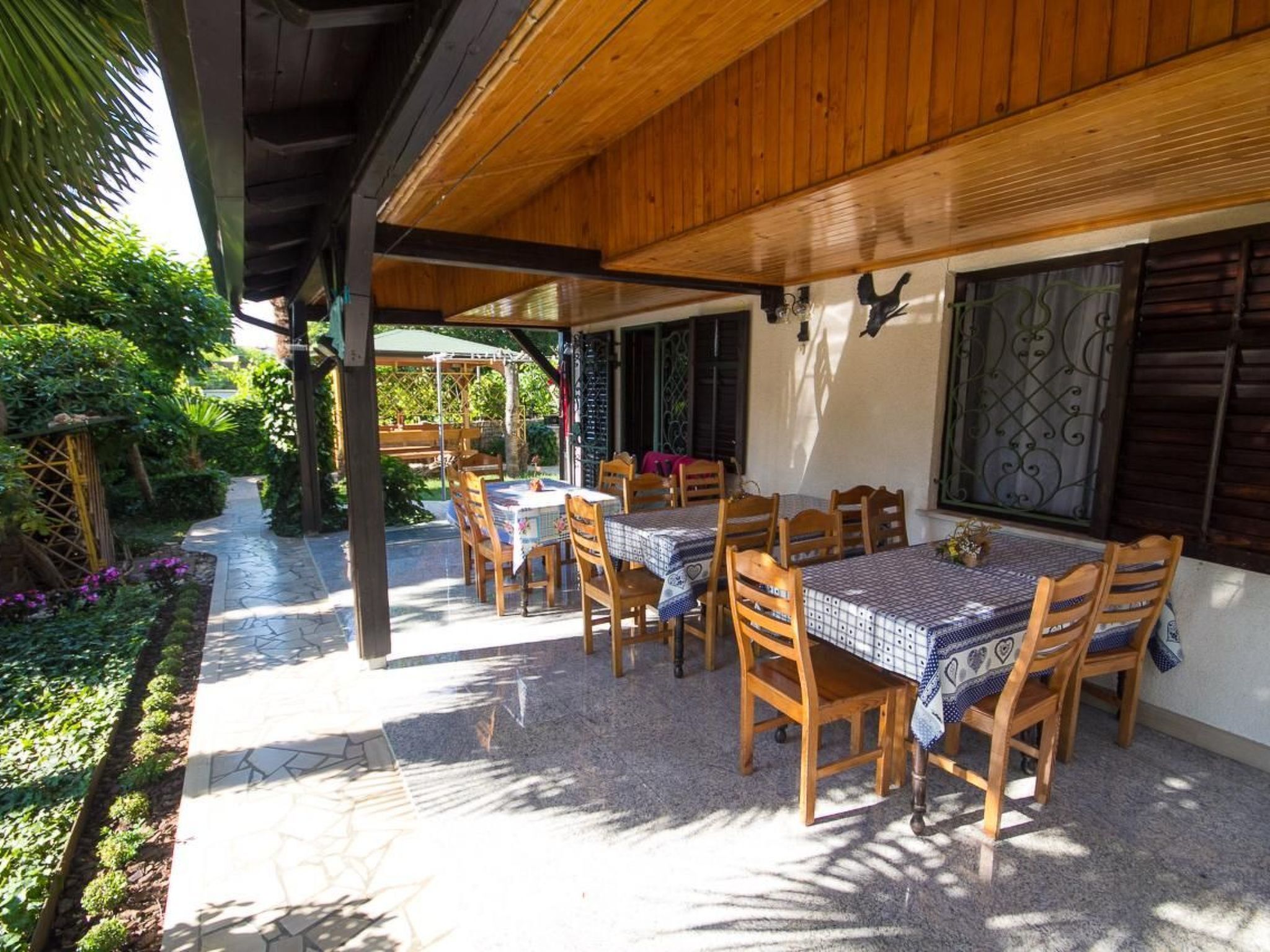 Um pátio externo com mesas e cadeiras de madeira, coberto por um telhado de madeira, cercado por vegetação e um caminho de pedra.