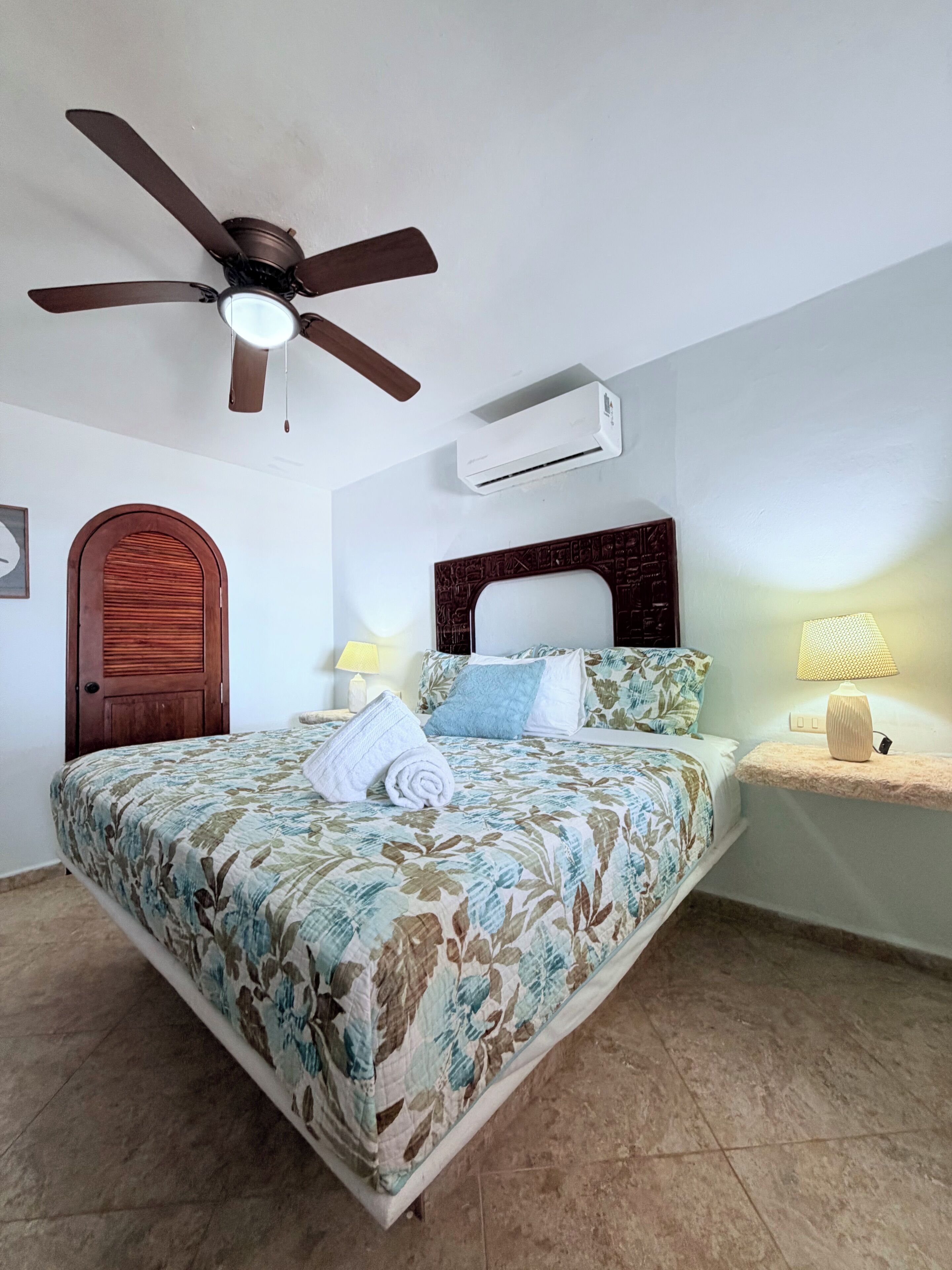 A bedroom with a bed, a ceiling fan, an air conditioning unit, a wooden door, and a nightstand with a lamp.
