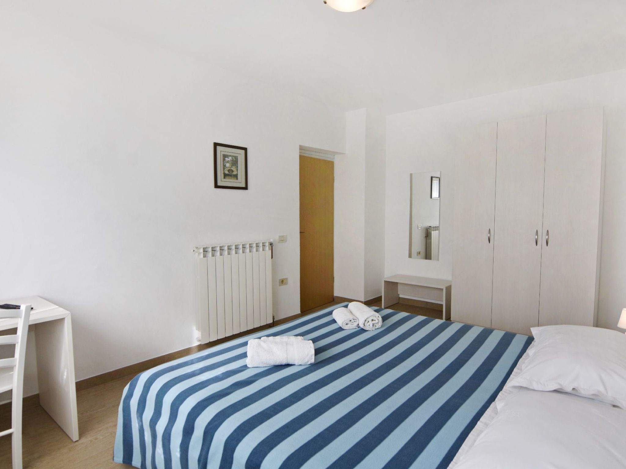 A hotel room with a bed, a desk, a radiator, a wardrobe, and a mirror.