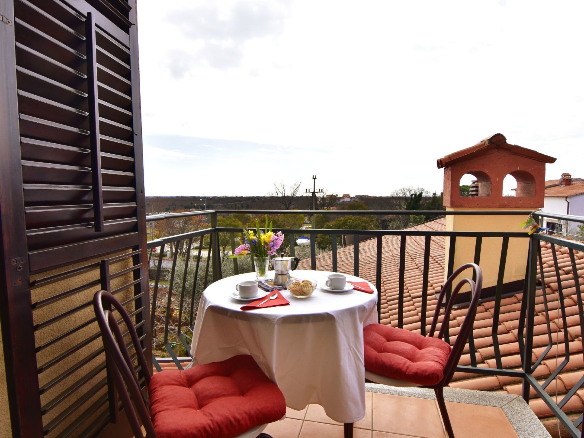 Un balcon avec une table dressée pour deux personnes, agrémenté d’une nappe blanche, de coussins rouges et d’un vase rempli de fleurs.
